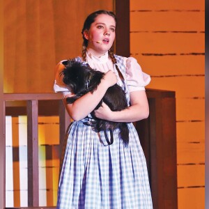 Kaida Collard, portraying Dorothy Gale, sings “Somewhere Over the Rainbow” while holding Toto (Annie Fry).